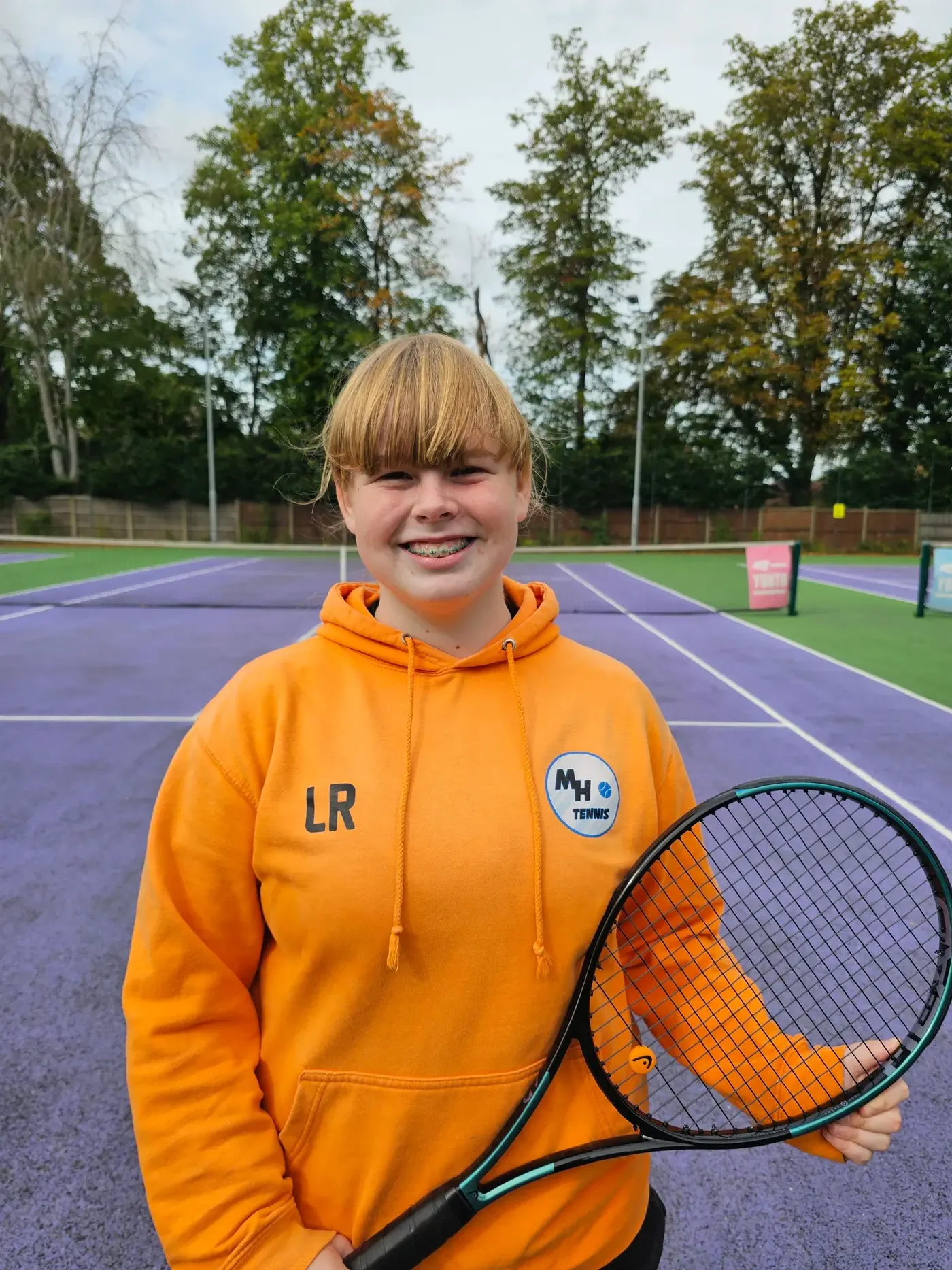 Lola, MH Tennis apprentice coach, smiling with a tennis racket on an outdoor tennis court